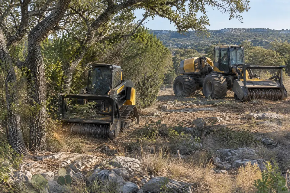 Lot clearing: compact track mulcher between oaks beside a larger articulated mulcher
