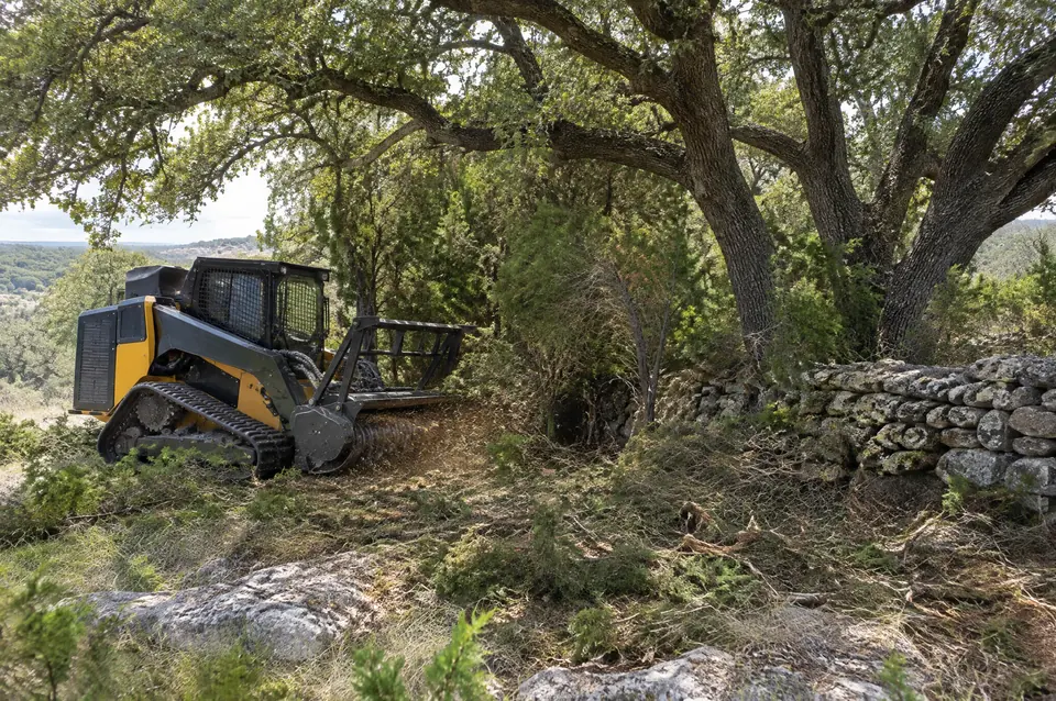 Forestry mulching near Bulverde: compact track mulcher clearing cedar under live oaks beside a dry-stack rock wall in Bulverde