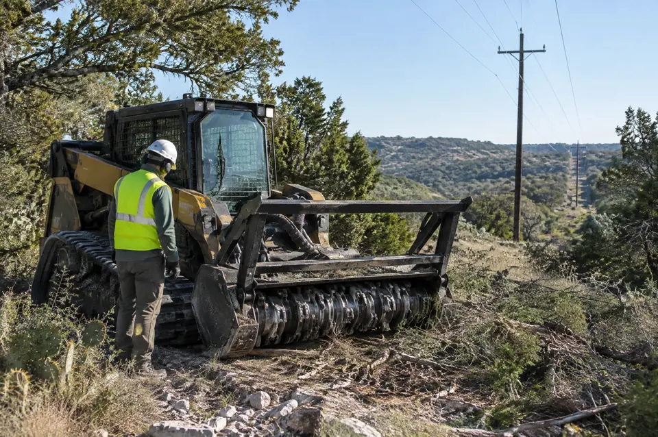 Easement clearing: crew member in hard hat and hi-vis vest checking a mulcher near a power line
