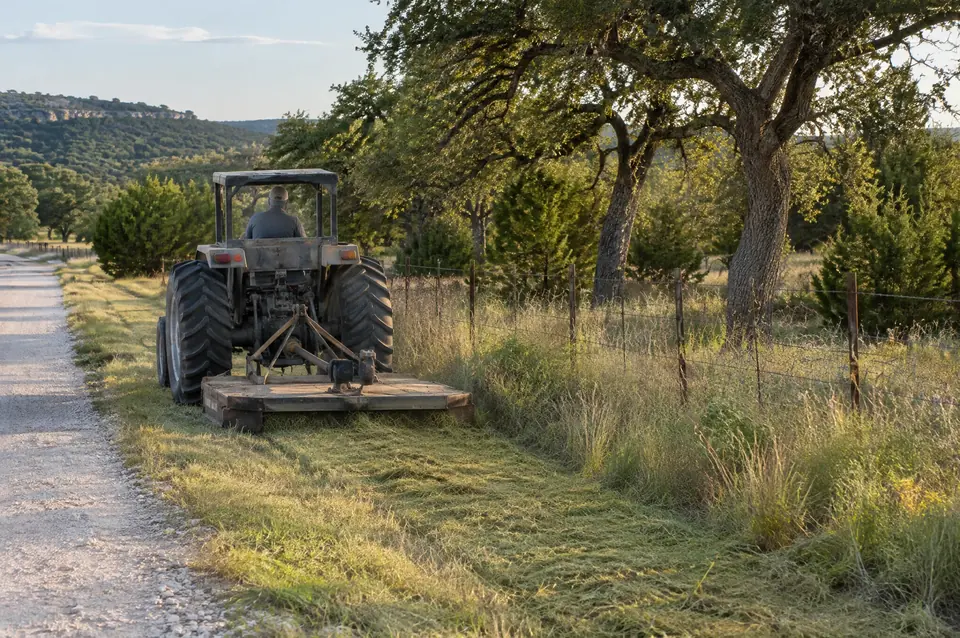 Grounds maintenance in Central Texas: gray utility tractor and shredder mowing a grassy strip beside a ranch road and fence