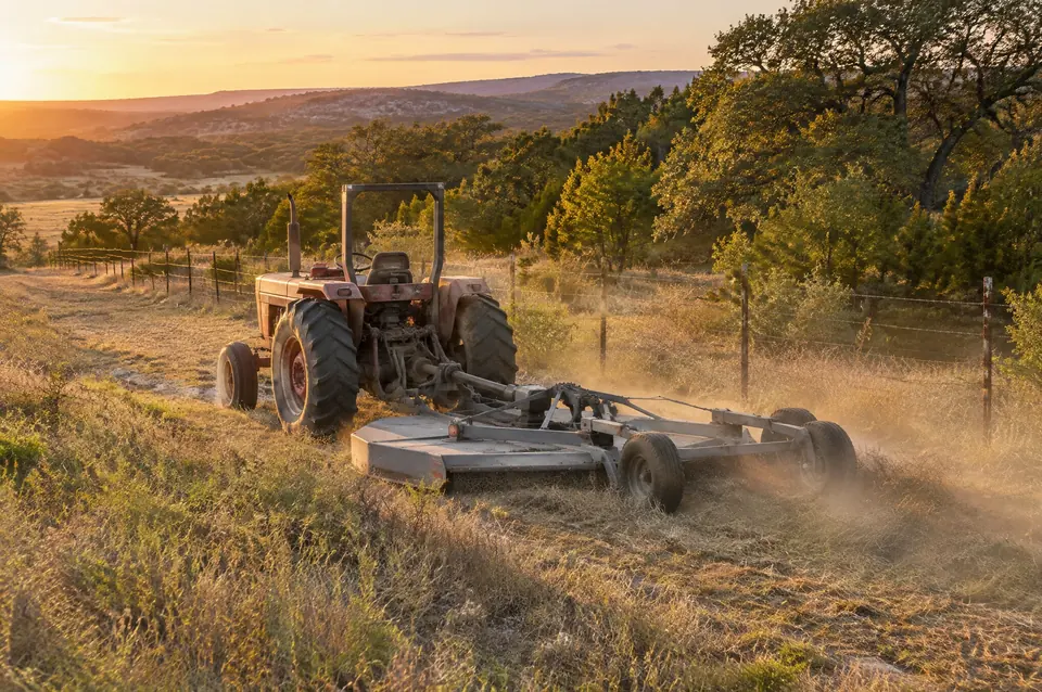 Land services in Central Texas: red tractor and batwing shredder mowing a field along a fence line at sunset