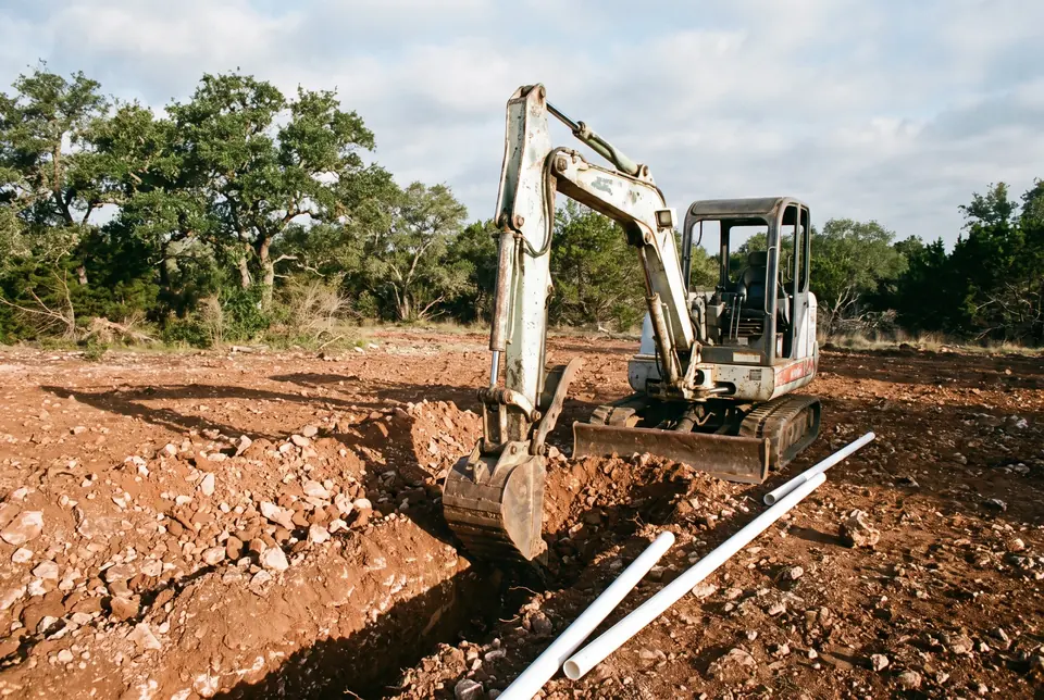 Site work and utility installation: excavator trenching rocky soil with waterline pipe laid beside the trench