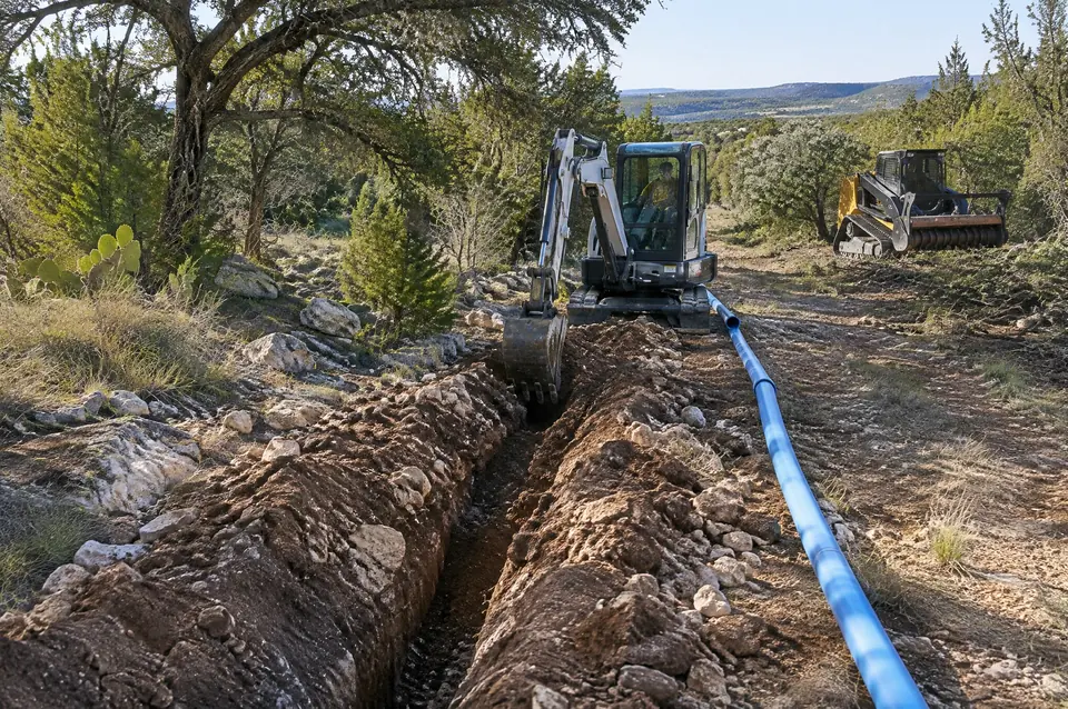 Dirt work: small excavator trenching rocky soil with blue waterline pipe alongside