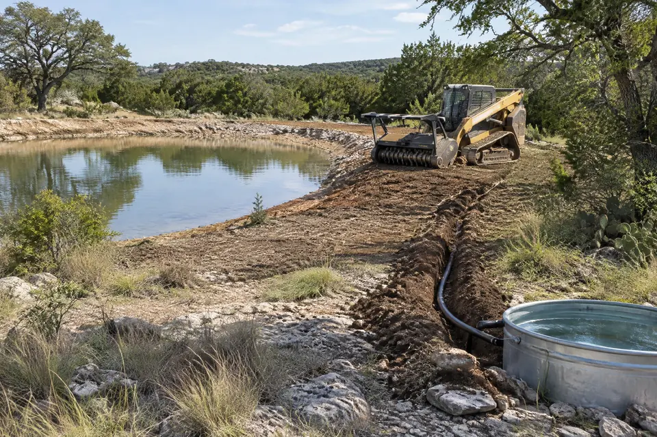 Fence line clearing: stock tank with a cleared bank and a new waterline trench to a trough